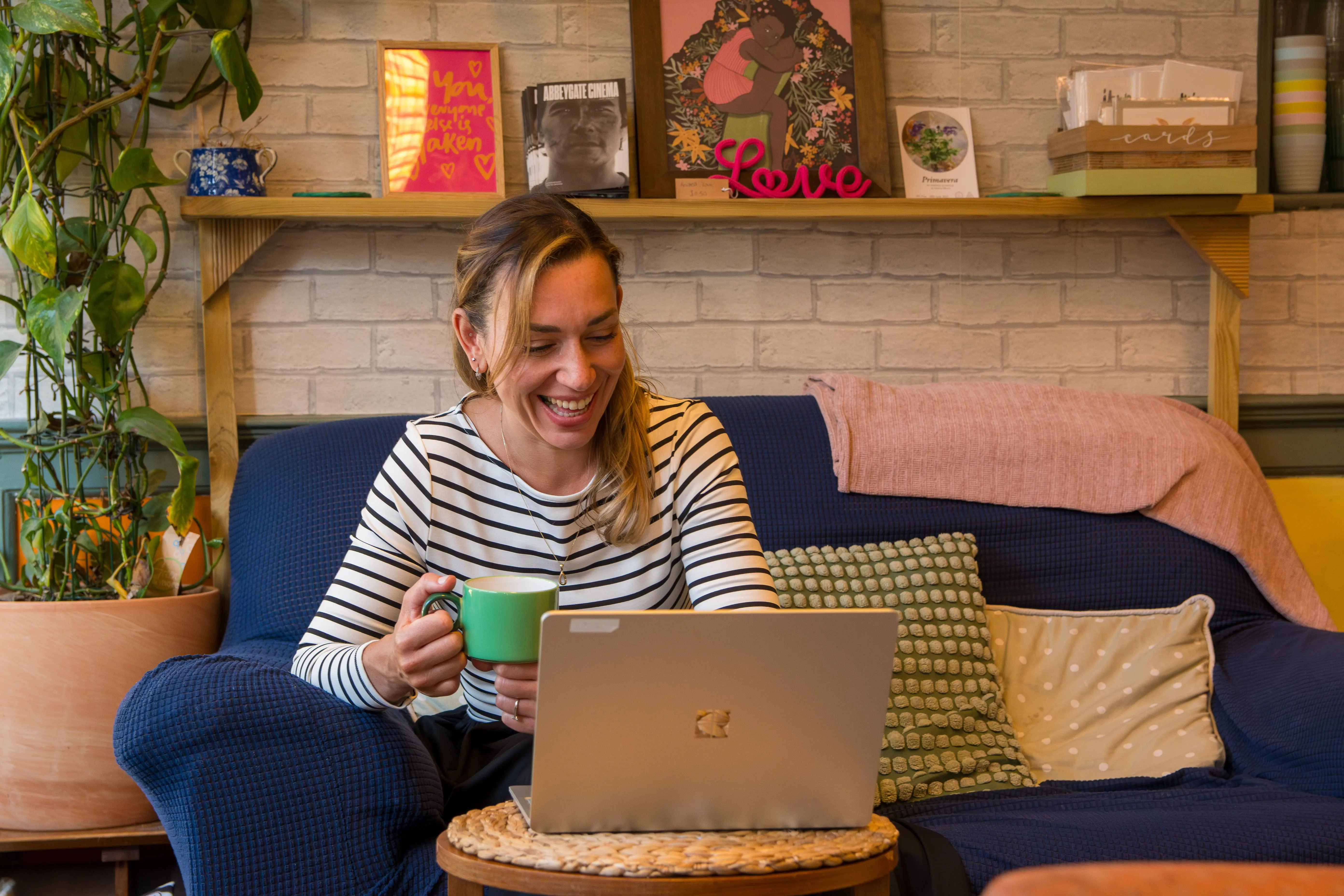 A female coach smiles while drinking coffee while on a client call via her laptop